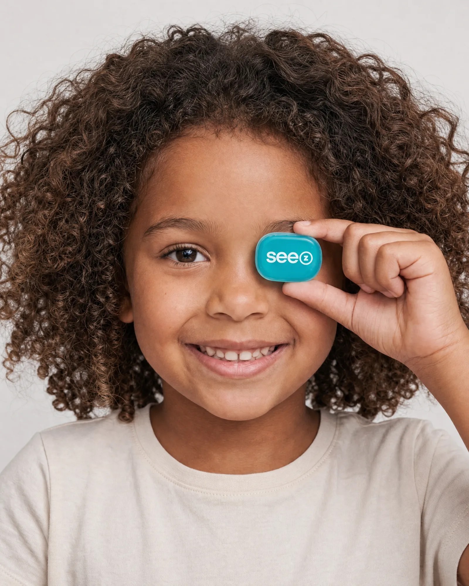 A smiling child holding a See Z ortho-k lens vial over one eye